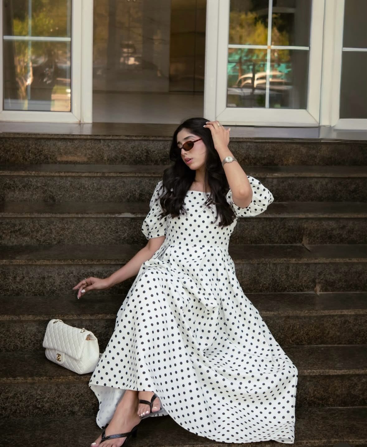 Woman in a black and white polka dot midi dress sitting on stairs with a white handbag.