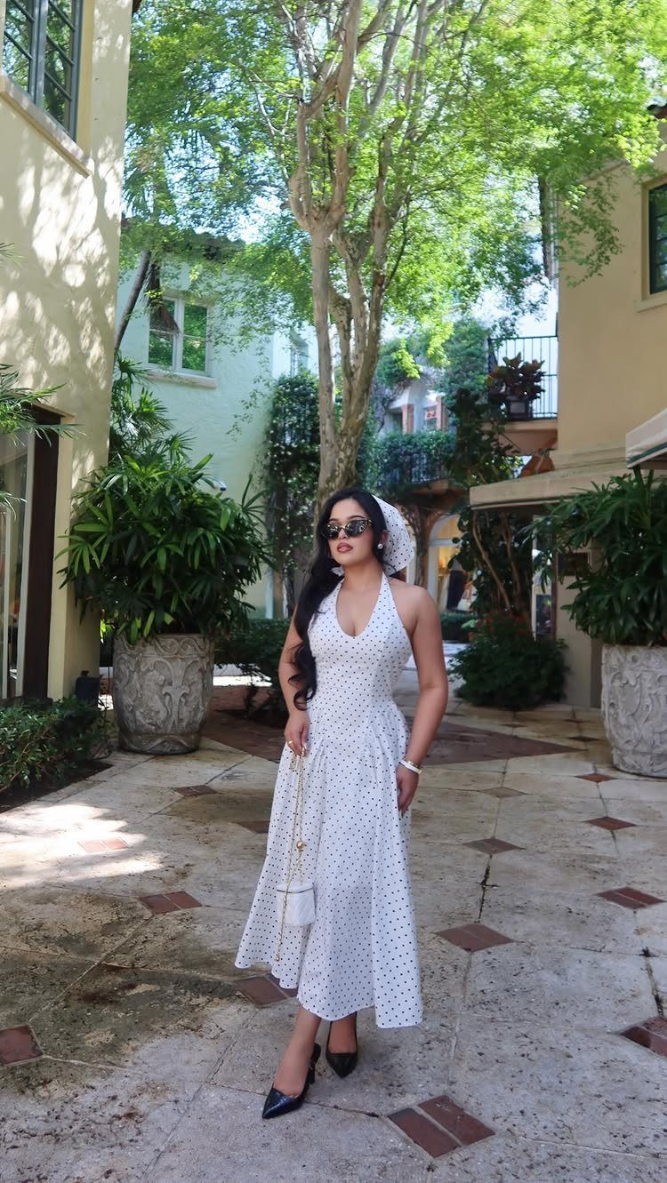 A woman posing in a white polka dot midi dress and black heels in a courtyard.