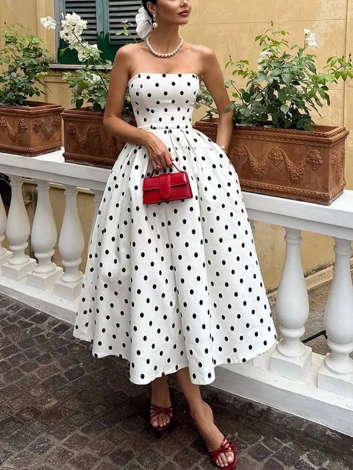 Woman in a white and black polka dot midi dress holding a red handbag.