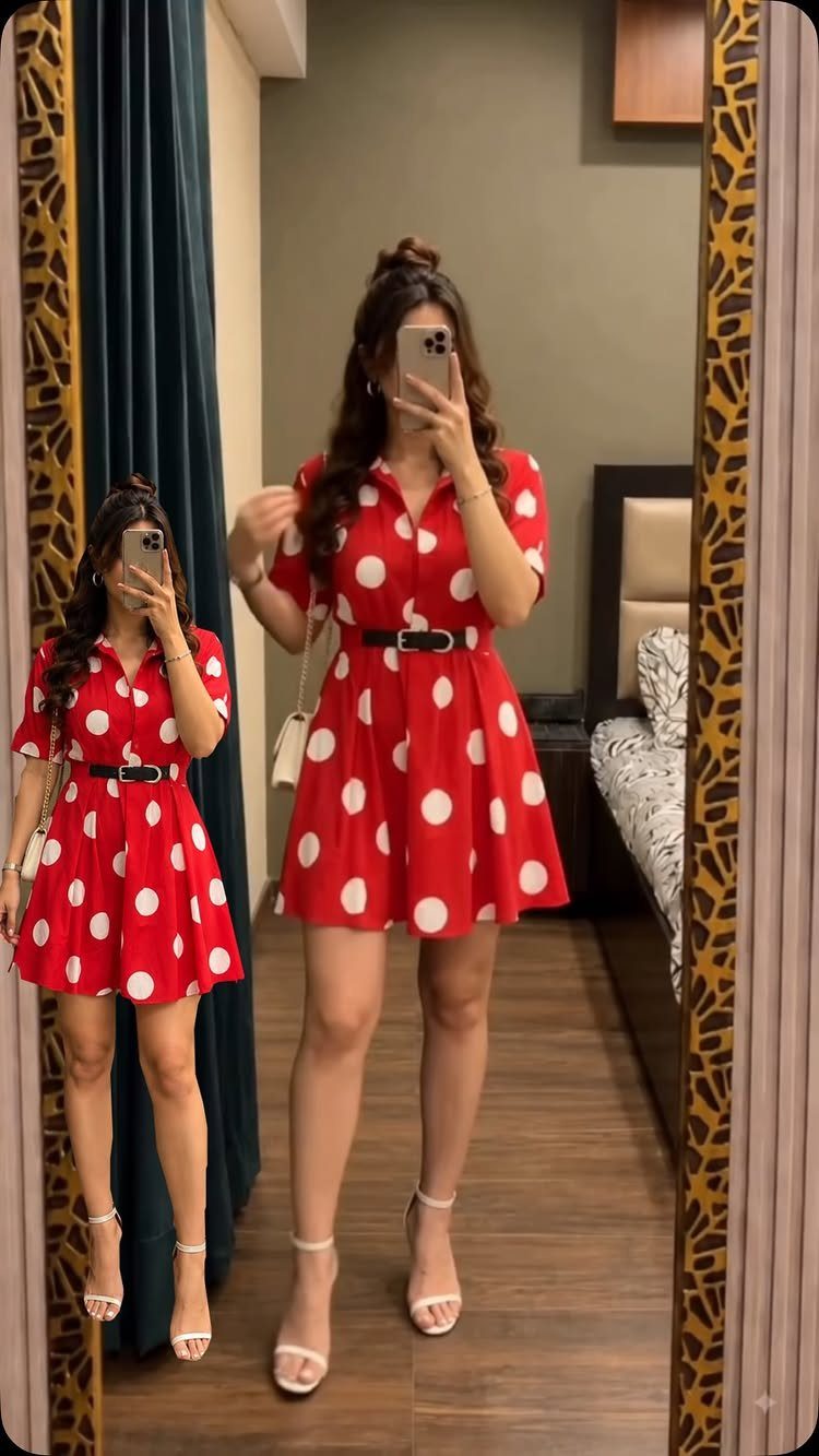 Woman in a red polka dot dress posing for a mirror selfie in a bedroom.