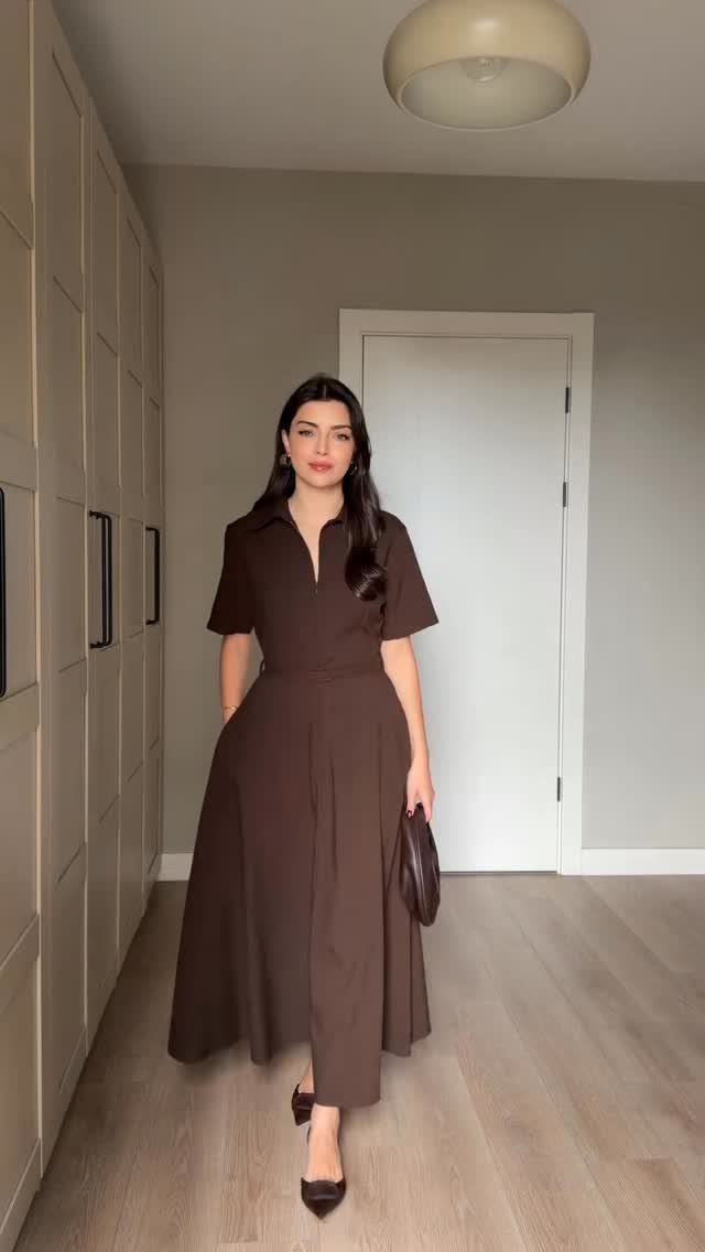 A woman wearing a cinched dark brown midi dress and holding a small handbag.