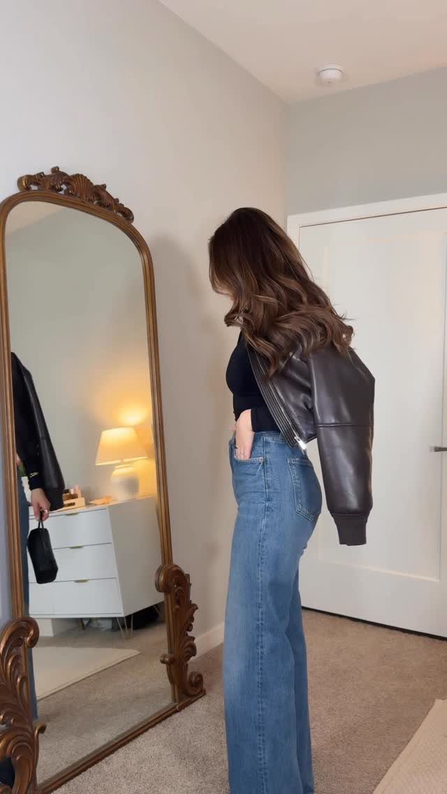 Woman wearing a black top, leather jacket, and blue wide-leg jeans standing near a mirror.