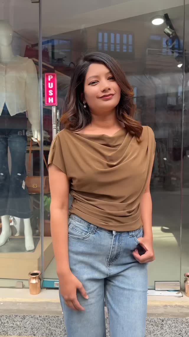 A woman wearing a brown cowl neck top and light blue jeans standing outdoors.