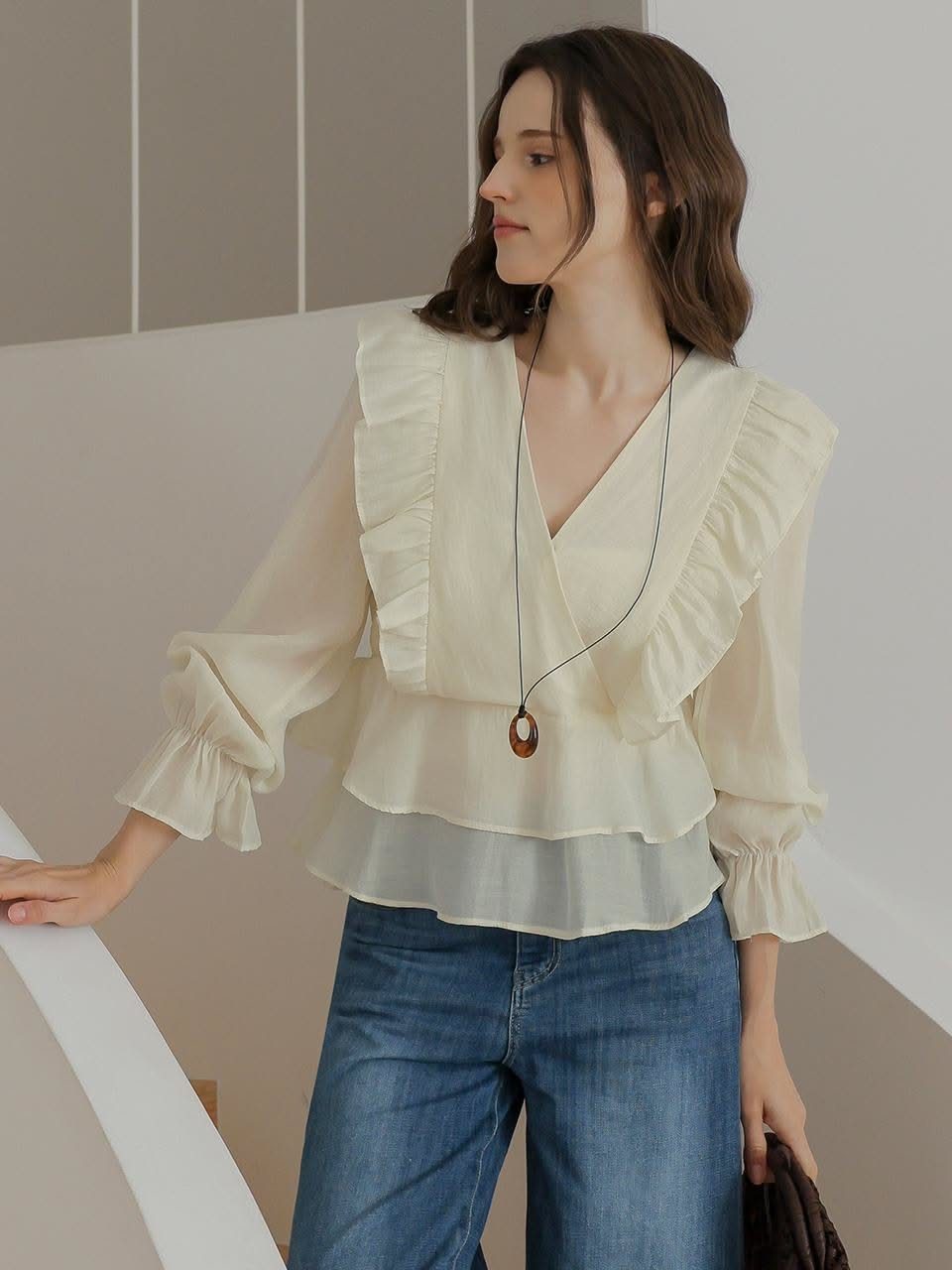 A woman wearing a cream ruffled blouse and blue jeans standing near a white wall.