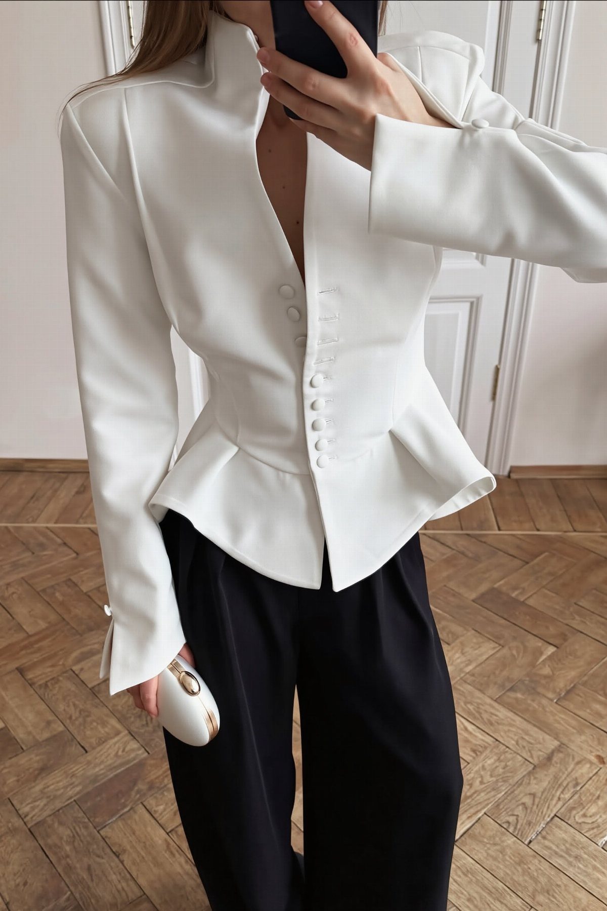 A person wearing a white structured peplum blazer and black trousers while taking a selfie.