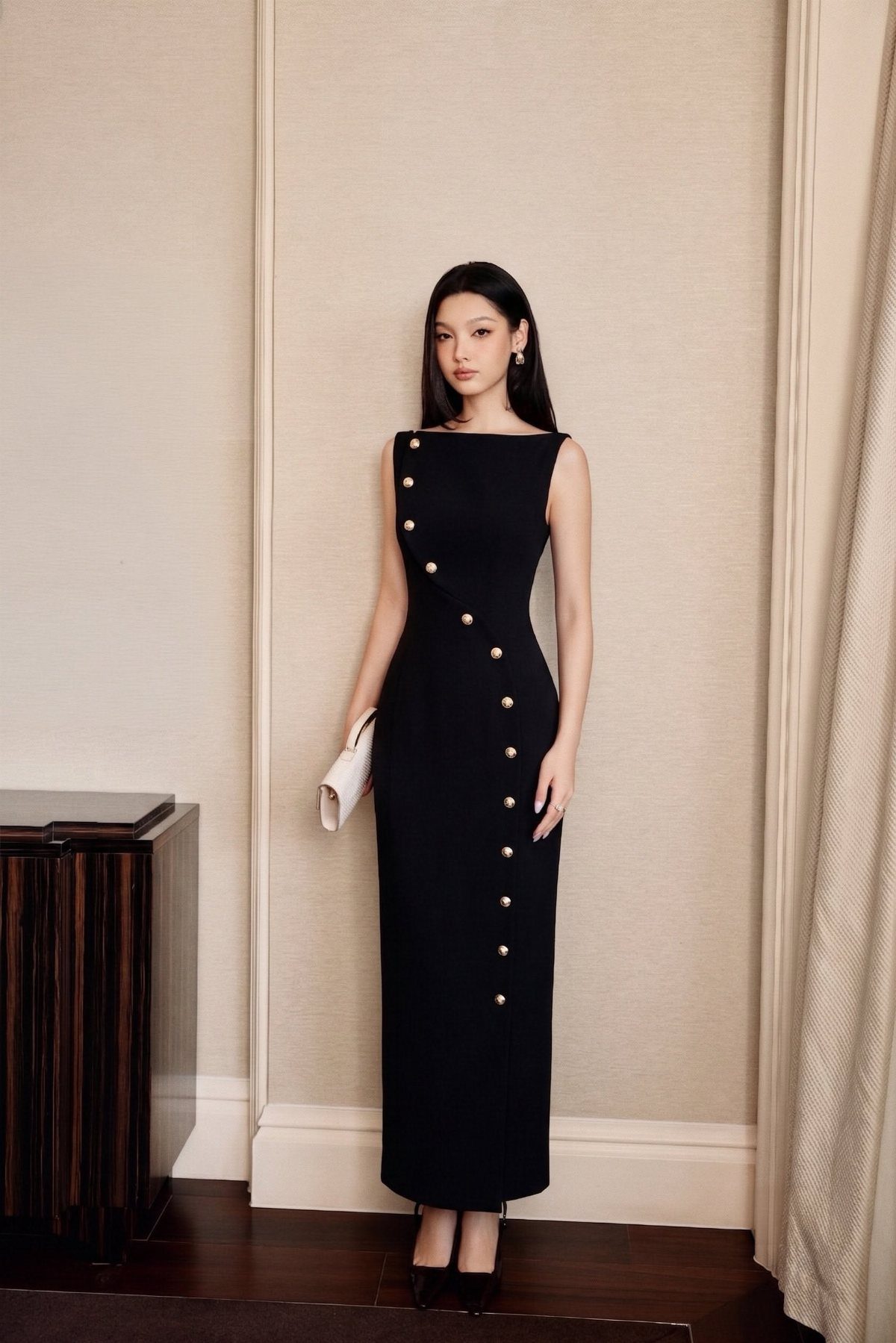 Woman in a long black structured dress with gold buttons standing against a neutral wall.