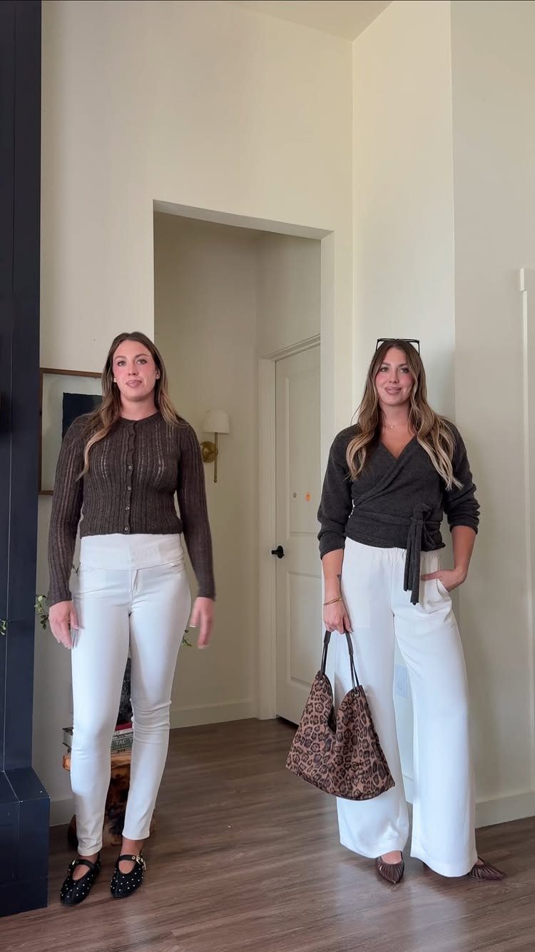 Two women posing in white trousers and dark tops inside a room.