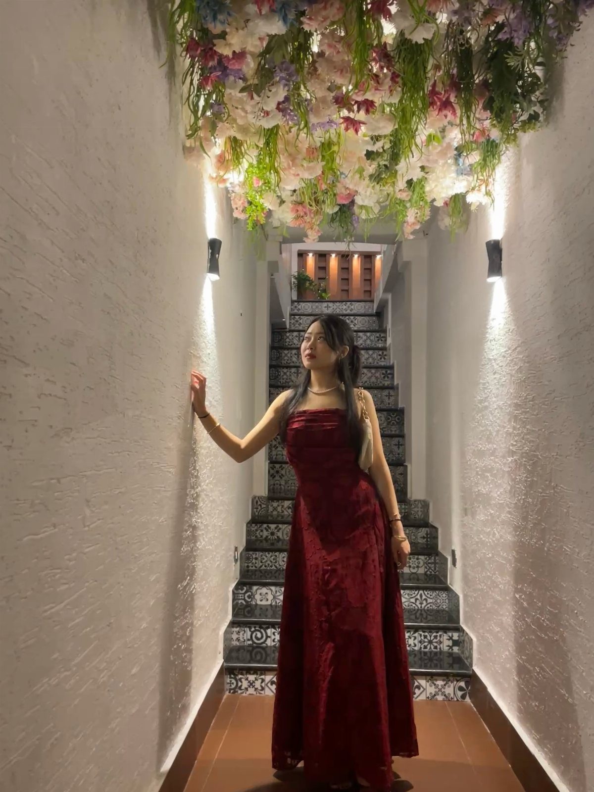 A woman posing in a long red velvet dress in a narrow hallway with floral decorations.