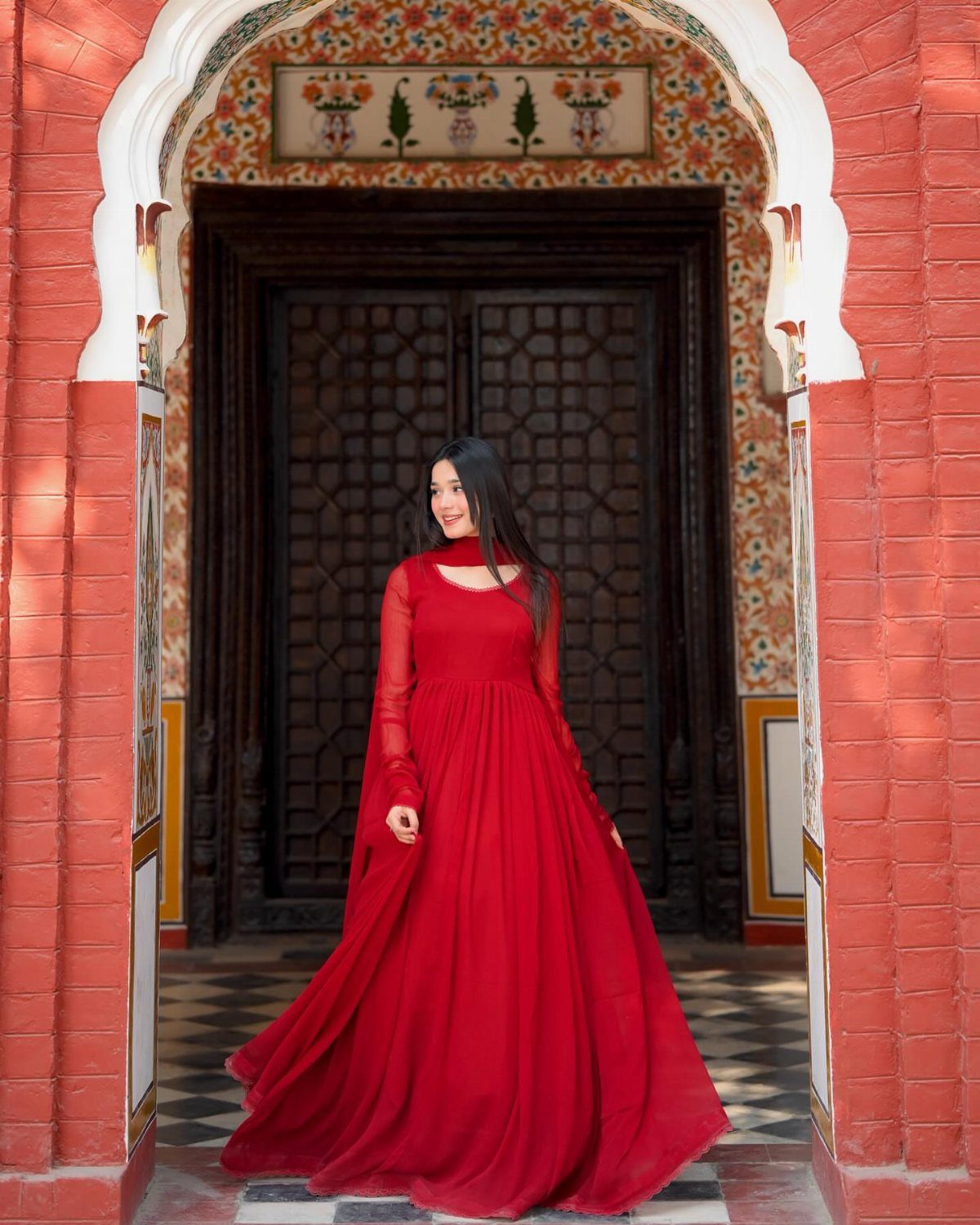 A woman in a flowing red chiffon dress standing in an ornate arched doorway.