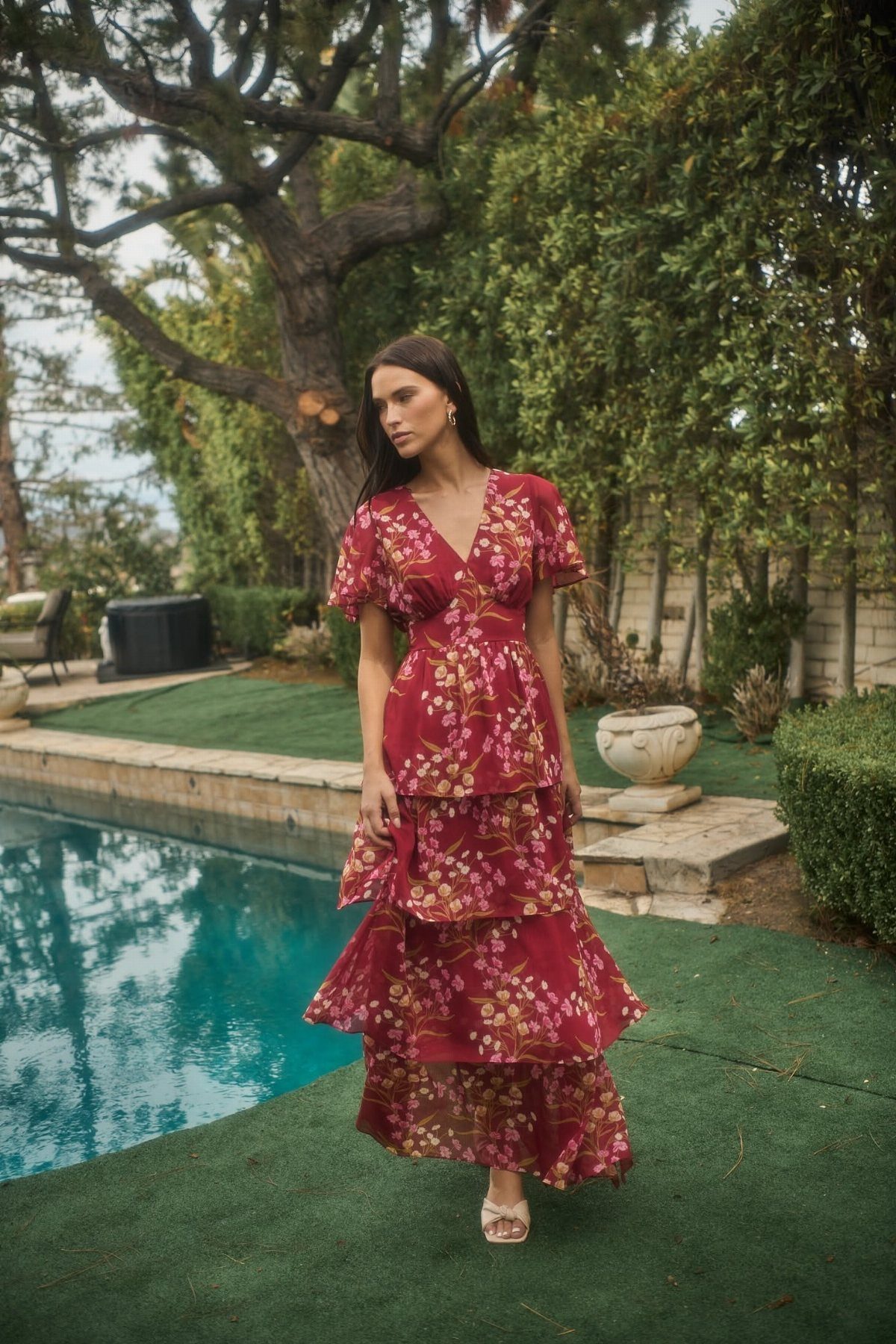 A woman wearing a long red floral patterned dress standing near a swimming pool.