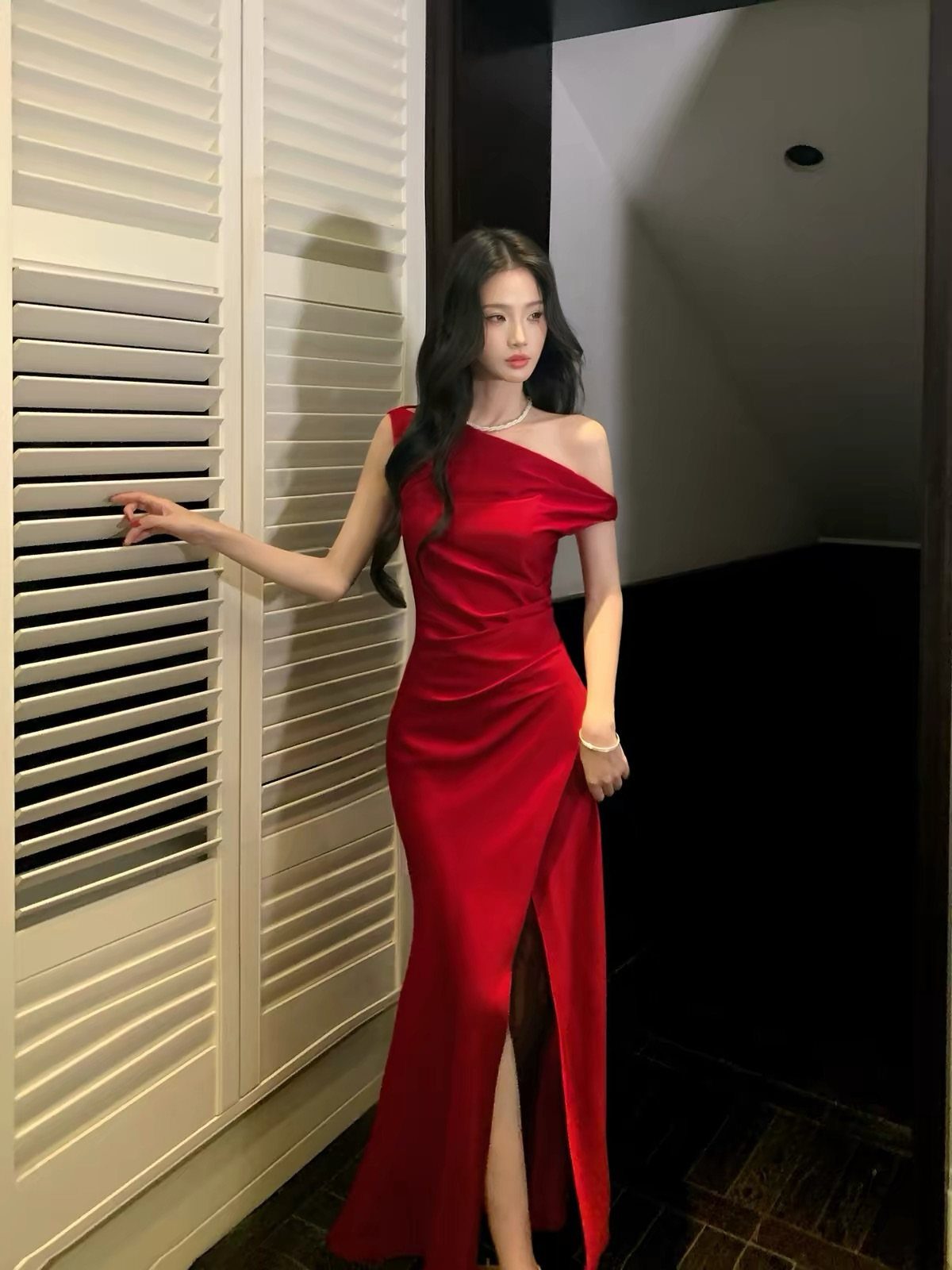 A woman in a red satin off-the-shoulder dress with a high thigh slit standing by white doors.