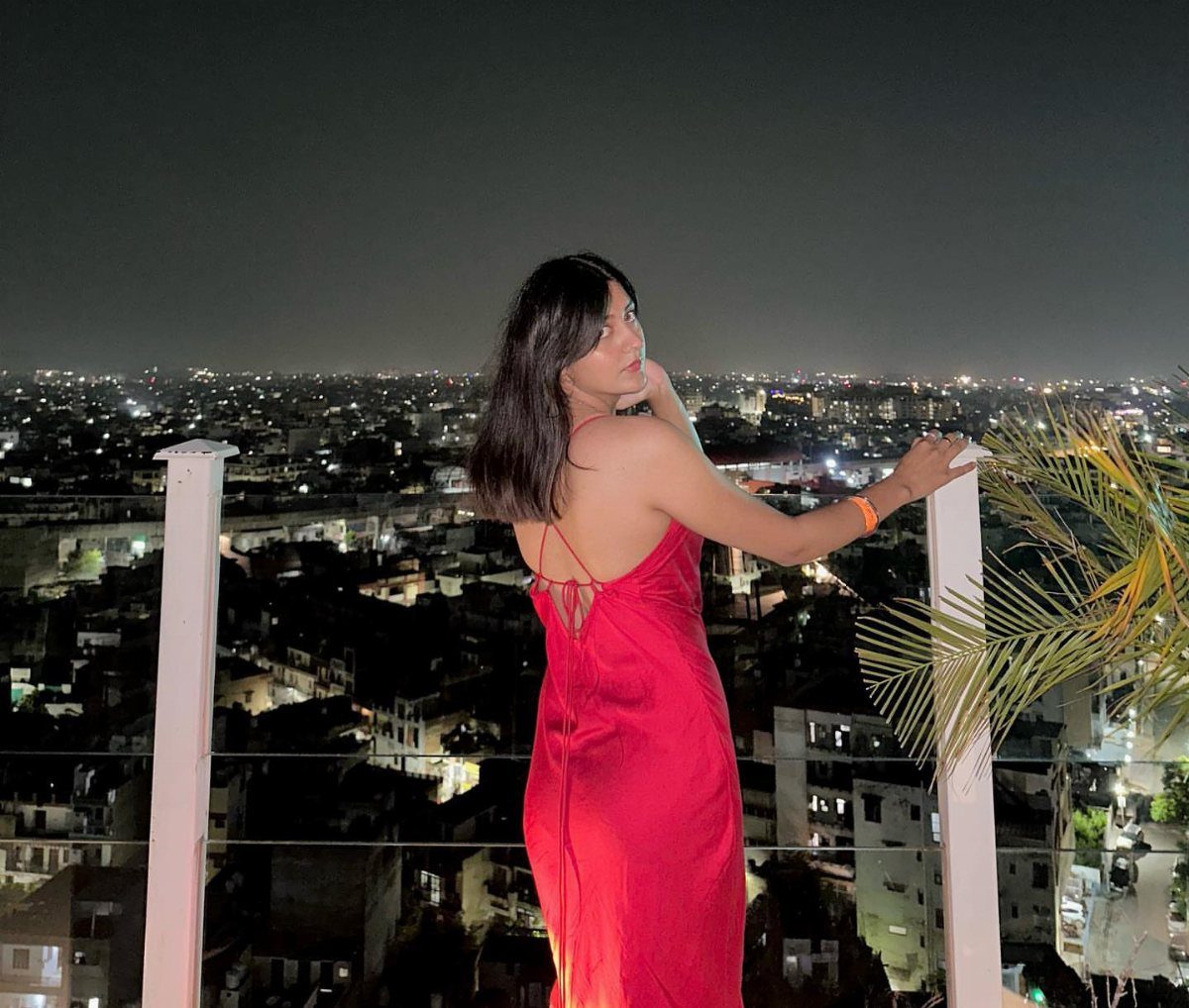 A woman in a red satin dress posing on a rooftop overlooking a city at night.