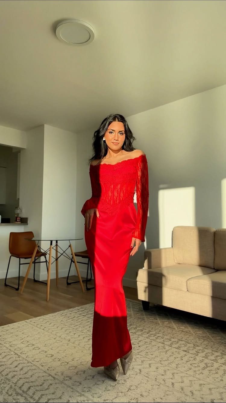 A woman posing in a long, formal red lace sleeve dress in a bright living room.