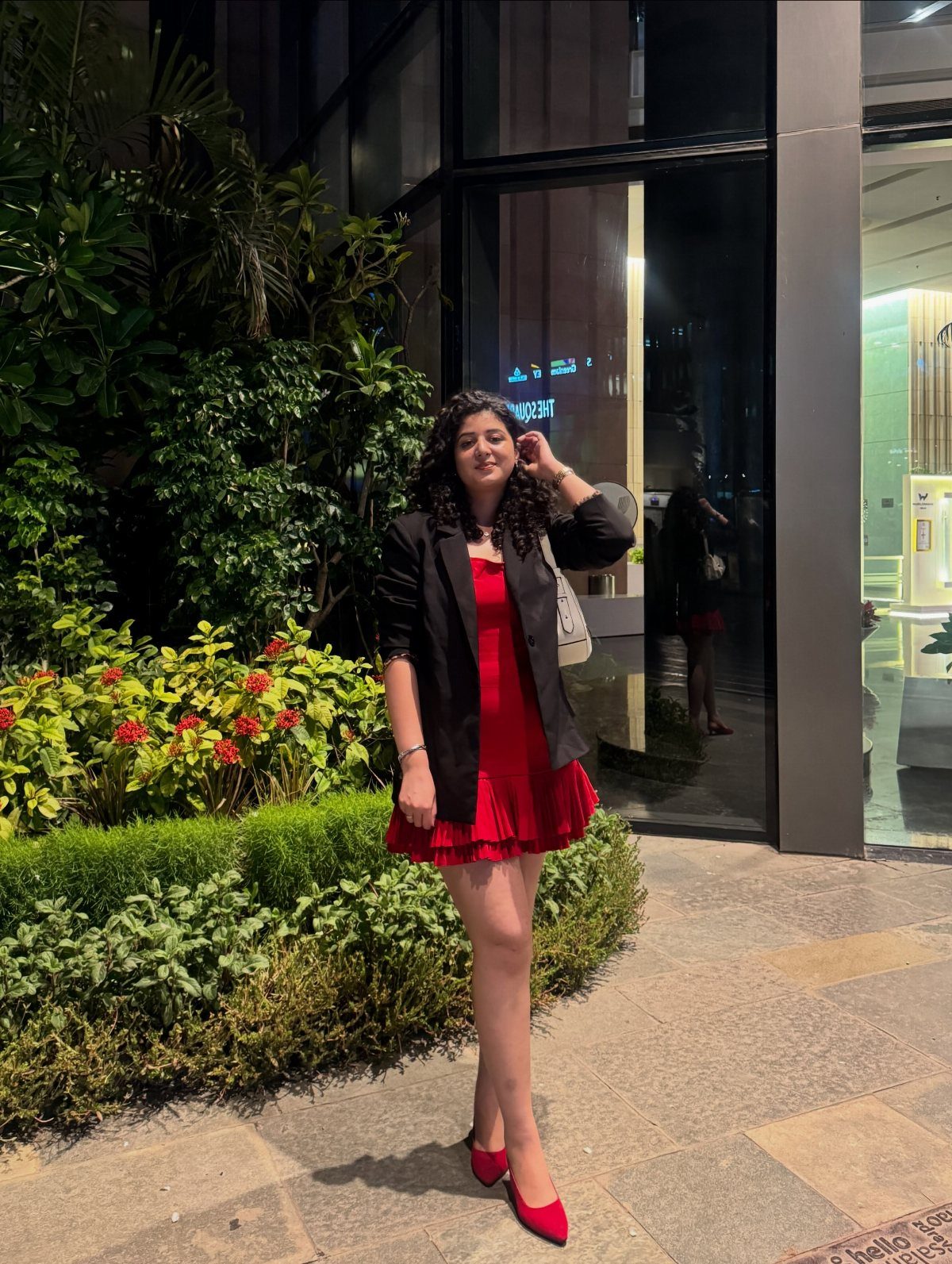 A woman posing in a red dress and black blazer next to green plants.
