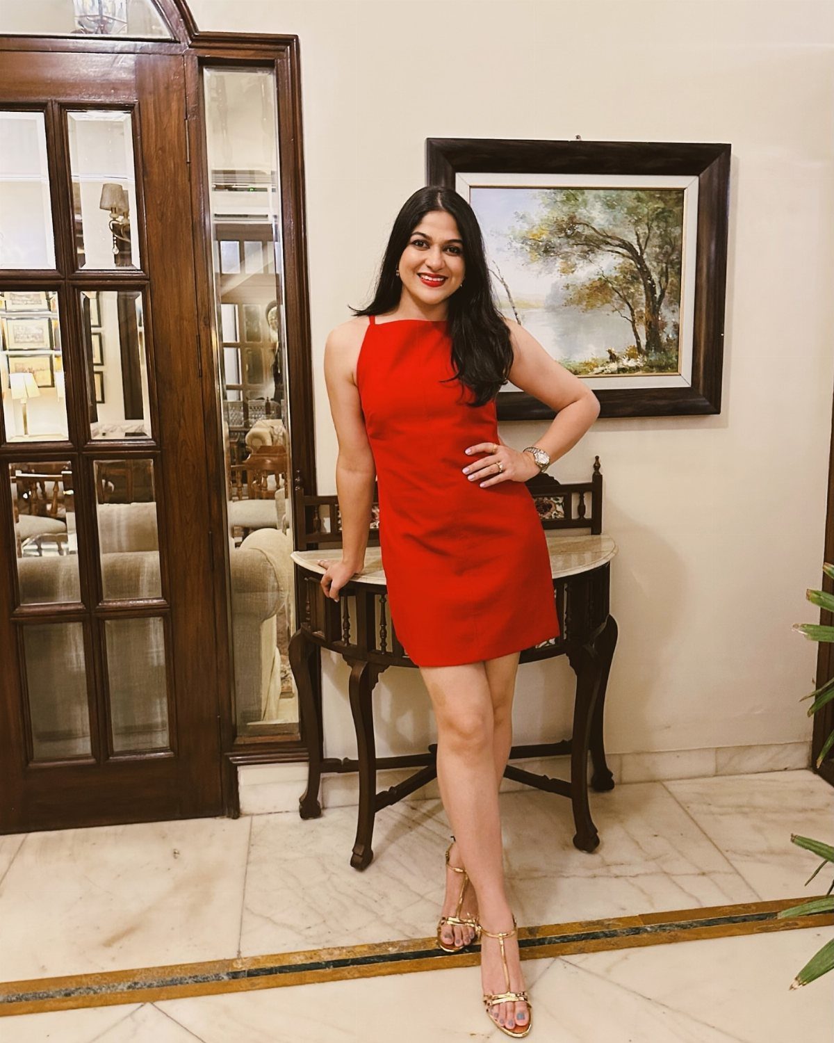A woman in a short red dress and gold metallic heels posing indoors.