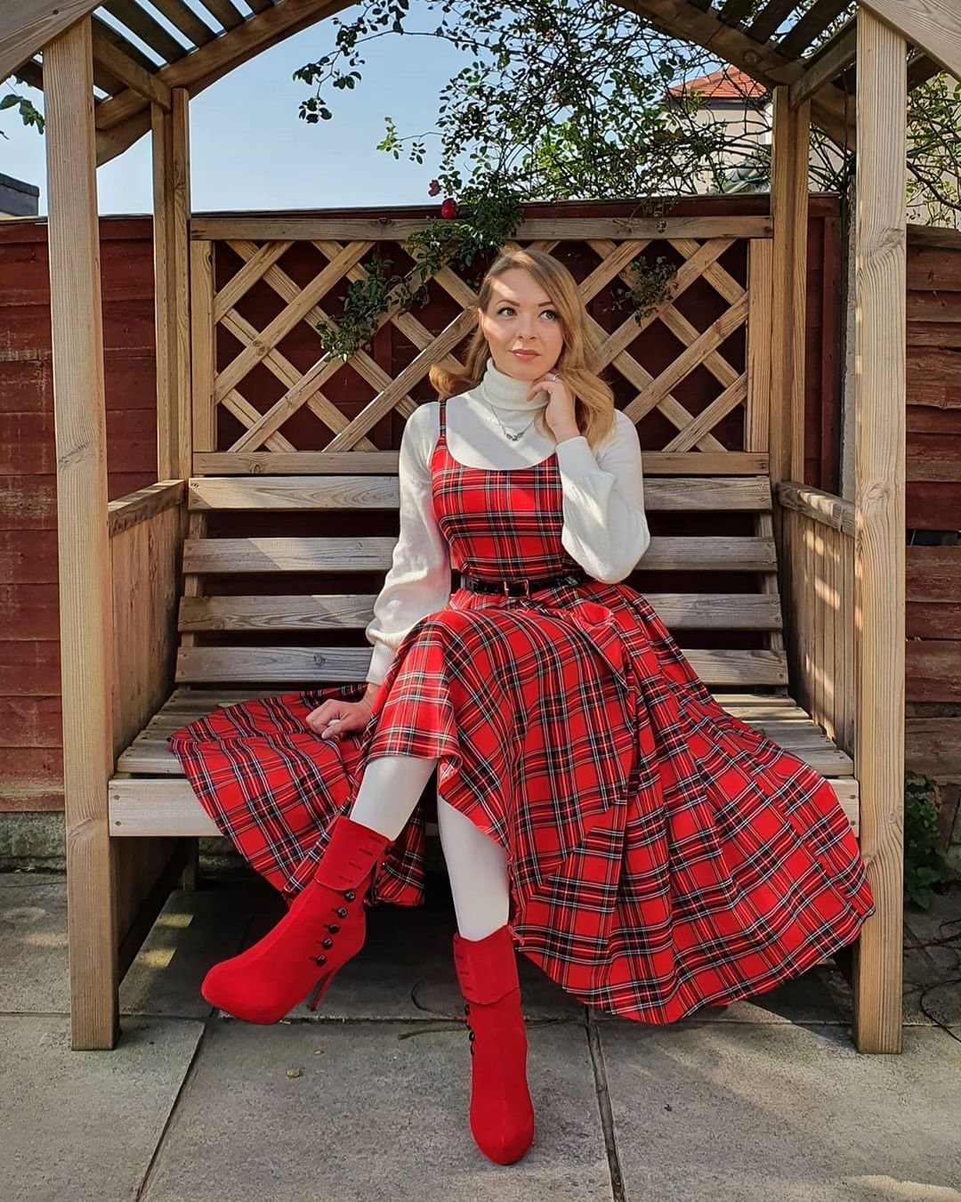 Woman in a red plaid high-low dress, white turtleneck, and red boots sitting on a wooden bench.