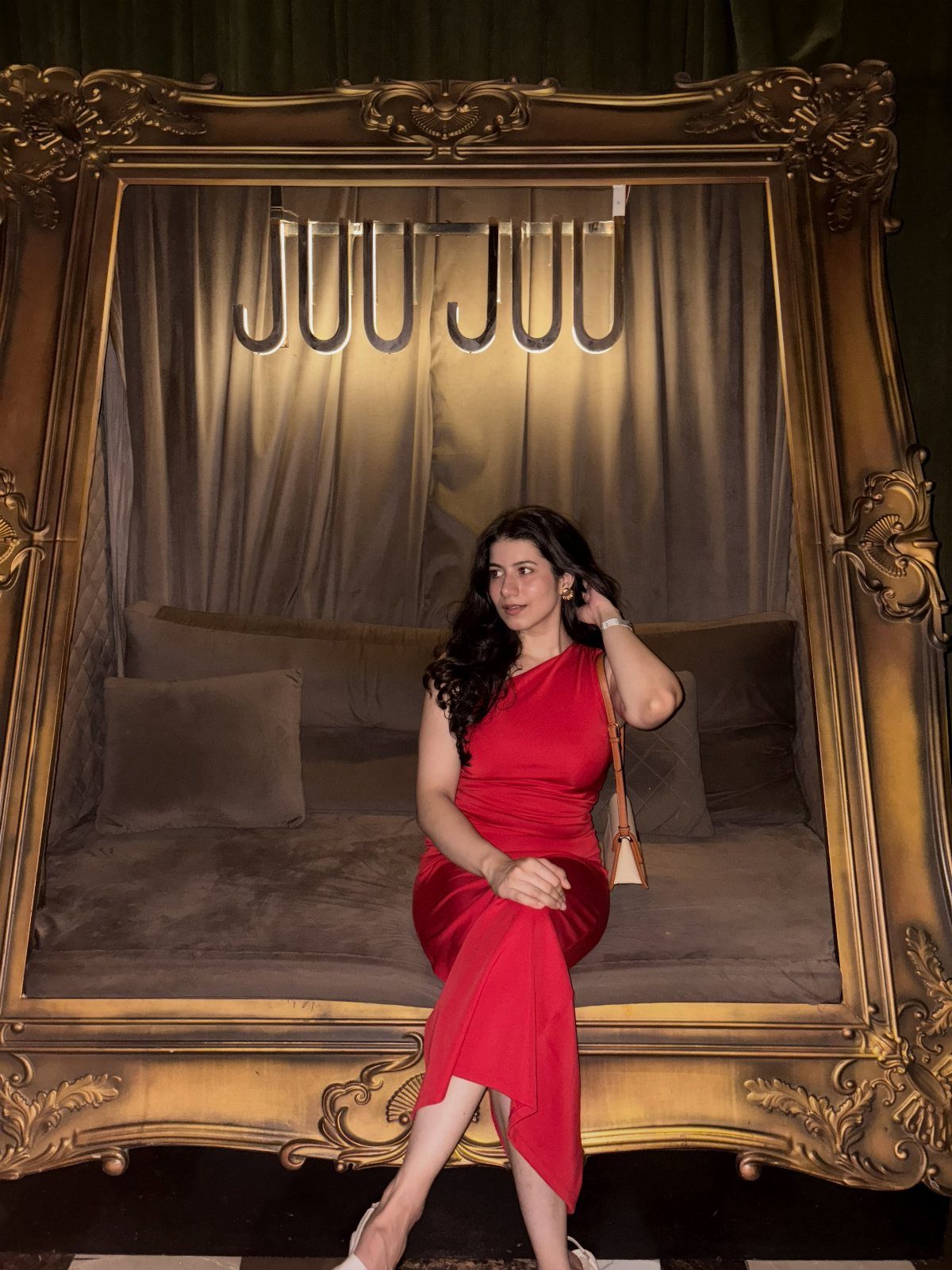 A woman in a red one-shoulder dress sitting inside a large gold frame.