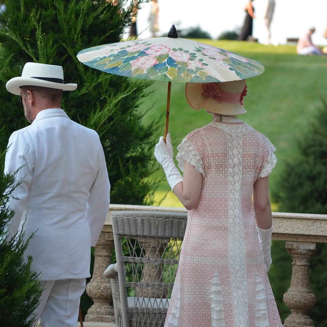 A woman in a pink dress holds a floral patterned parasol outdoors near a man in white.