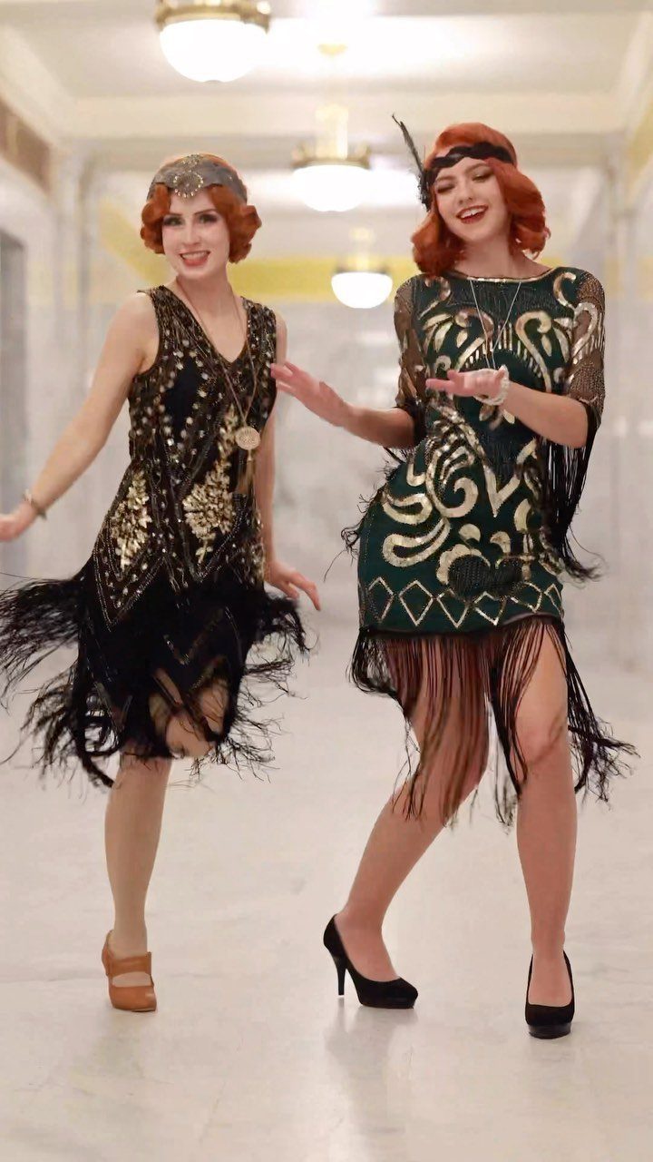 Two women wearing sequined and fringed flapper dresses dancing in a bright hallway.