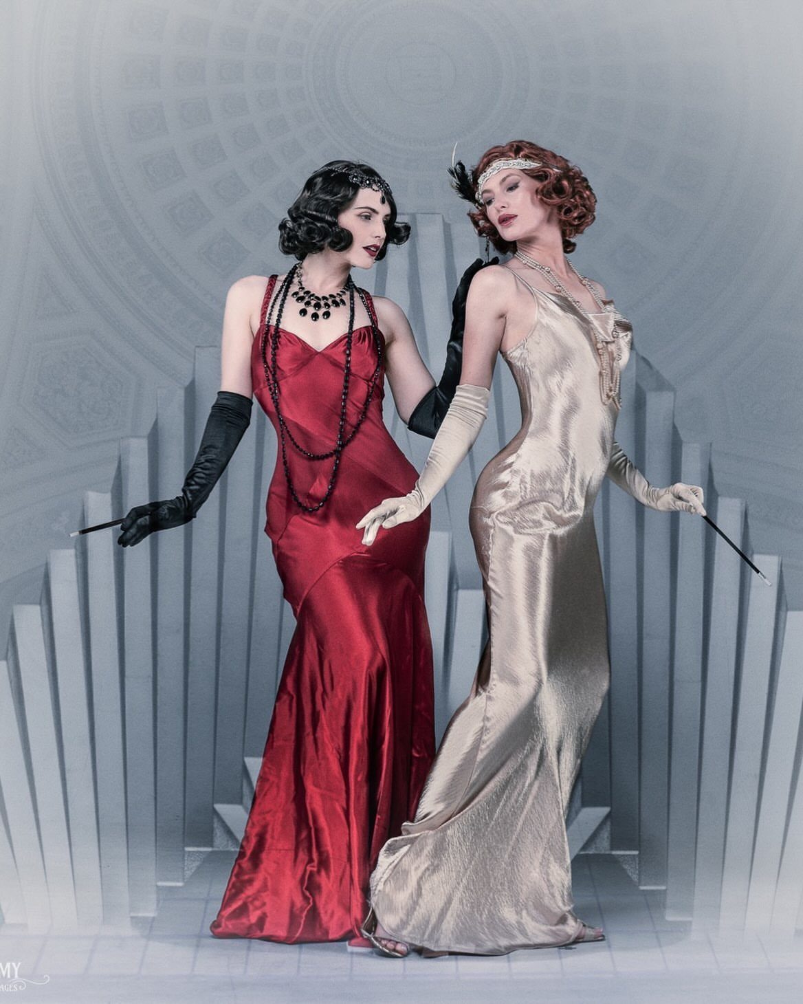 Two women posing in elegant, floor-length satin gowns with long gloves and vintage accessories.