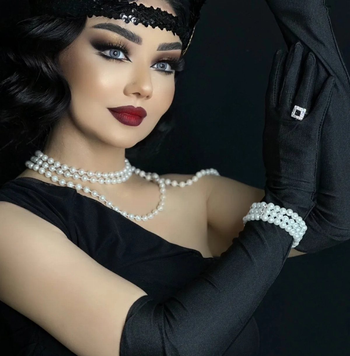 Woman with dark wavy hair wearing black satin gloves and long pearl necklaces.