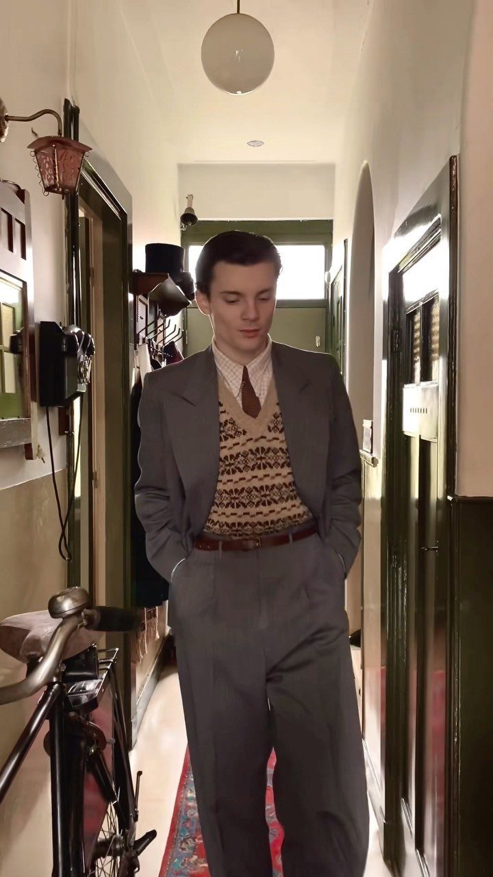 A man wearing a grey vintage-style suit and patterned sweater vest in a hallway.
