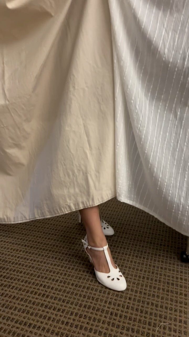 White T-strap ballet flats with decorative cutouts worn on a person's foot.