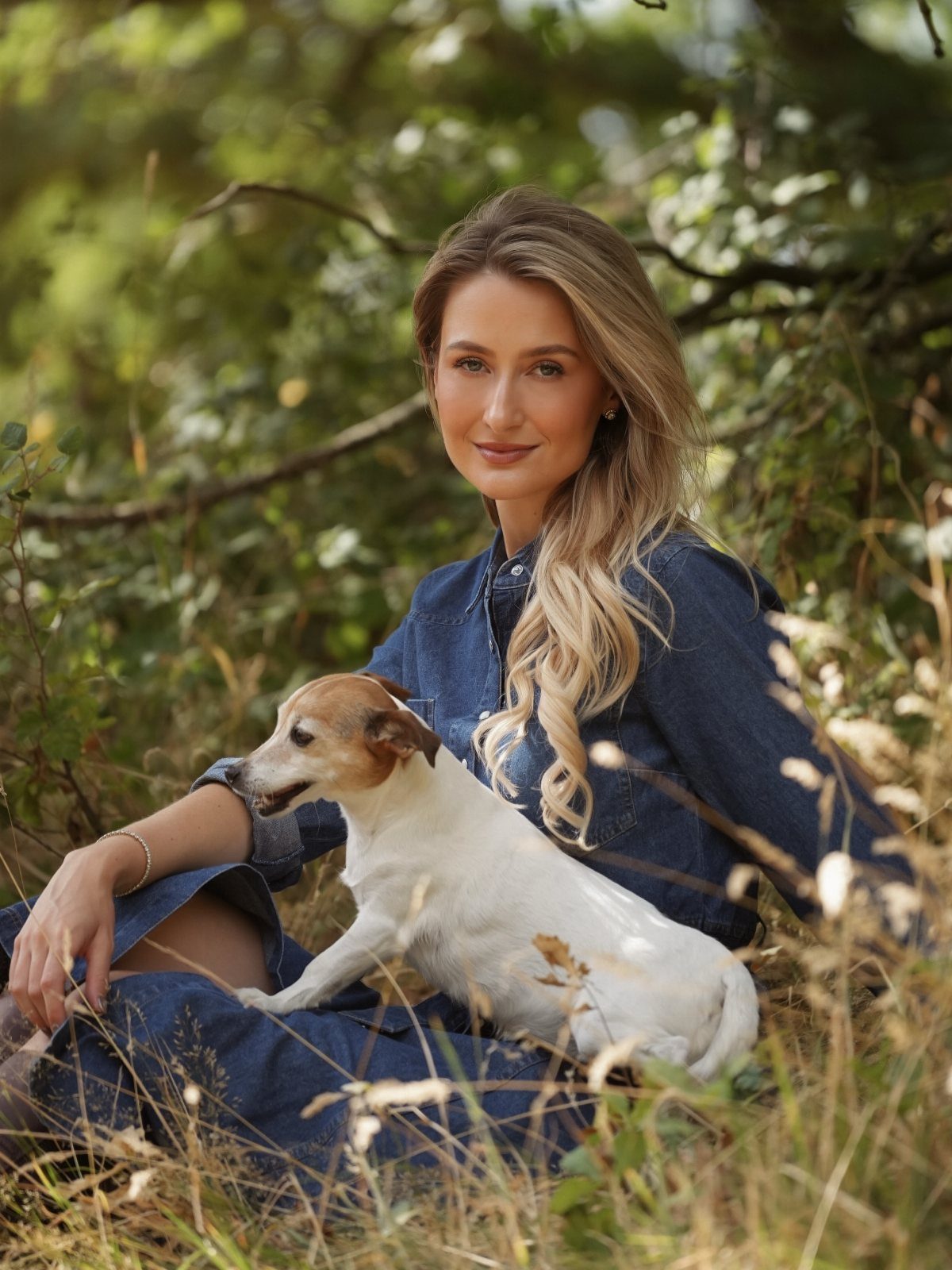 A woman in a blue denim dress sitting in the grass with a small dog.