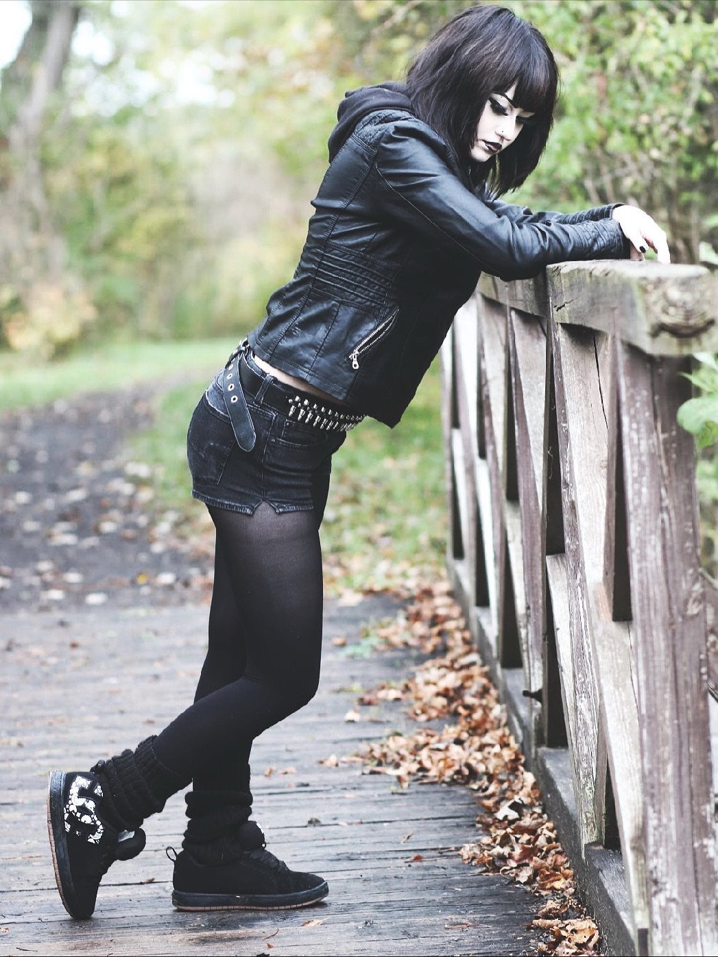 A young woman in an edgy black leather jacket and studded skirt poses on a wooden bridge.
