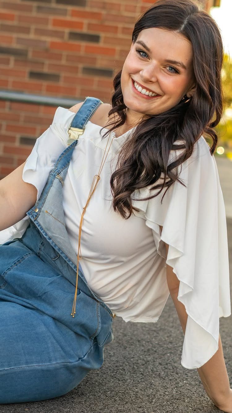Smiling young woman wearing a white ruffled blouse and blue denim overalls outdoors.
