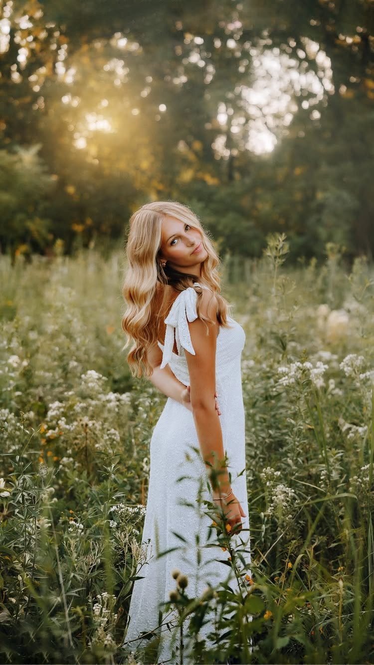 Blonde woman in a white dress posing in a sunlit field of wildflowers.