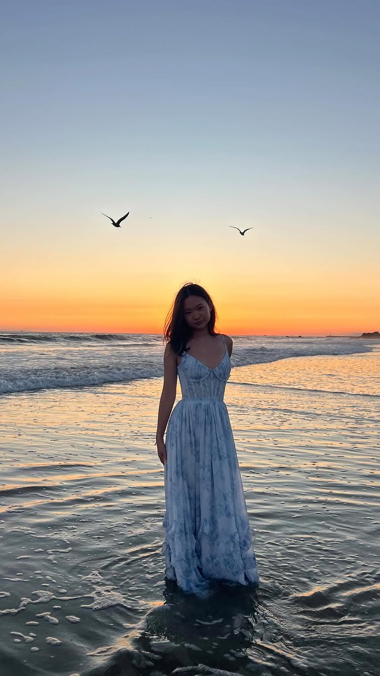 A young woman in a long flowy dress standing in ocean waves at sunset.