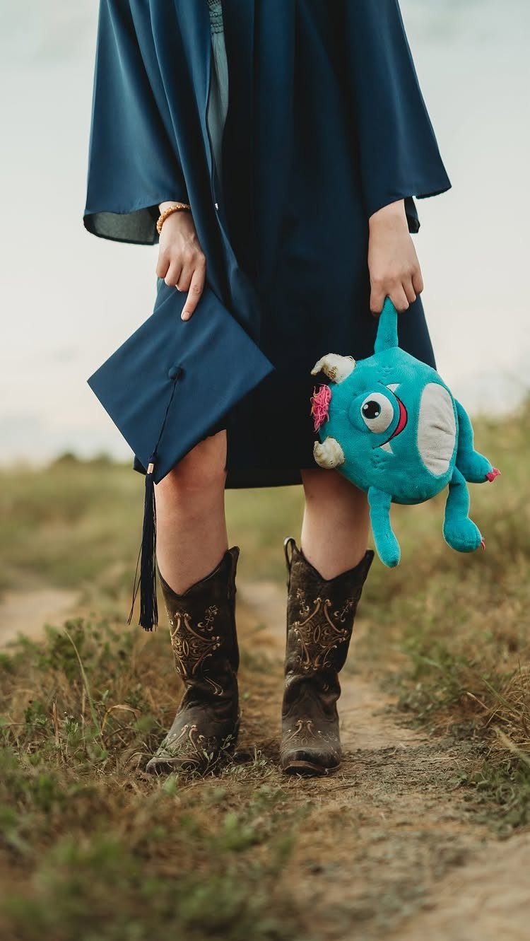 A graduate in a navy gown holding a cap and blue plush toy while wearing cowboy boots.