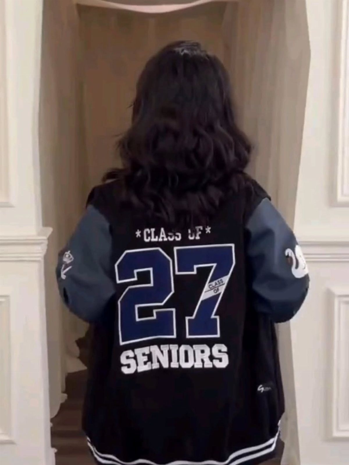 Person wearing a black and grey varsity jacket with Class of 27 Seniors printed on the back.