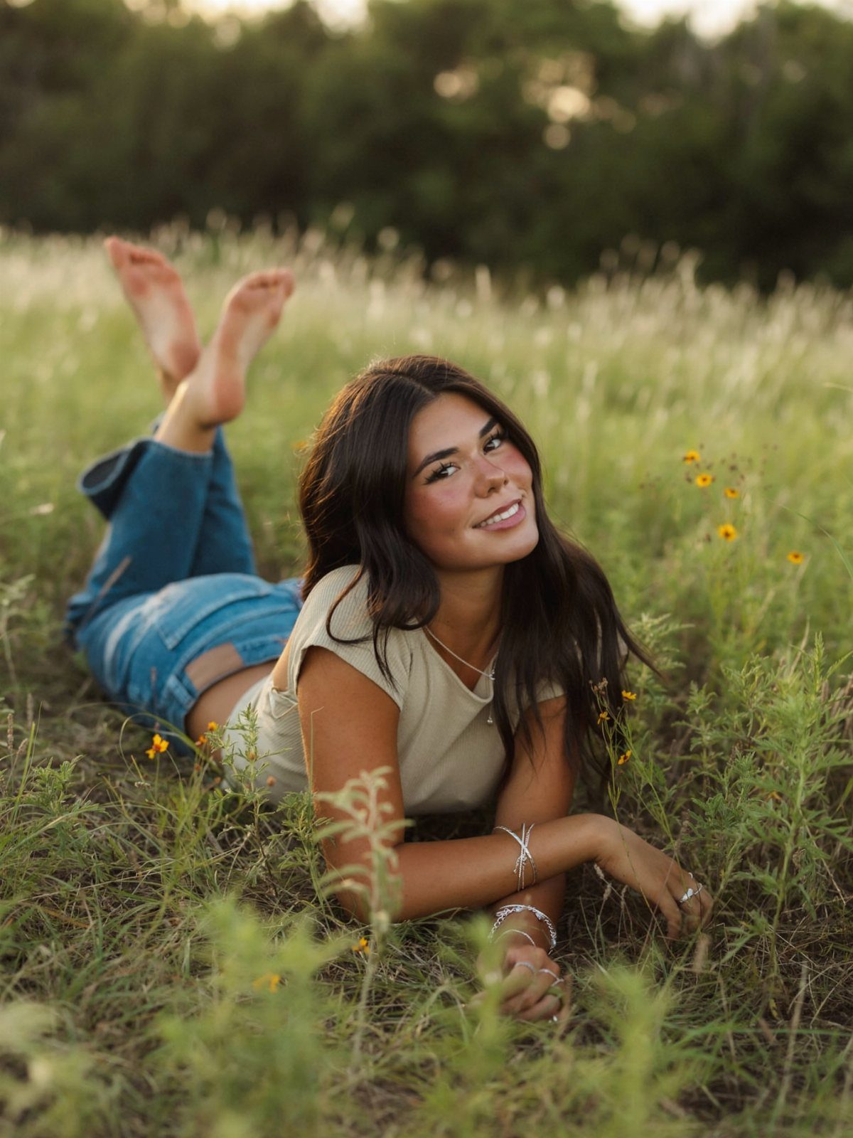 A smiling young woman with long dark hair lying in a grassy field during sunset.