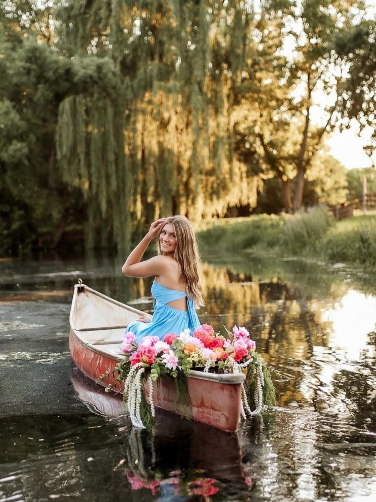 A smiling woman in a blue dress sitting in a flower-decorated canoe on a lake.