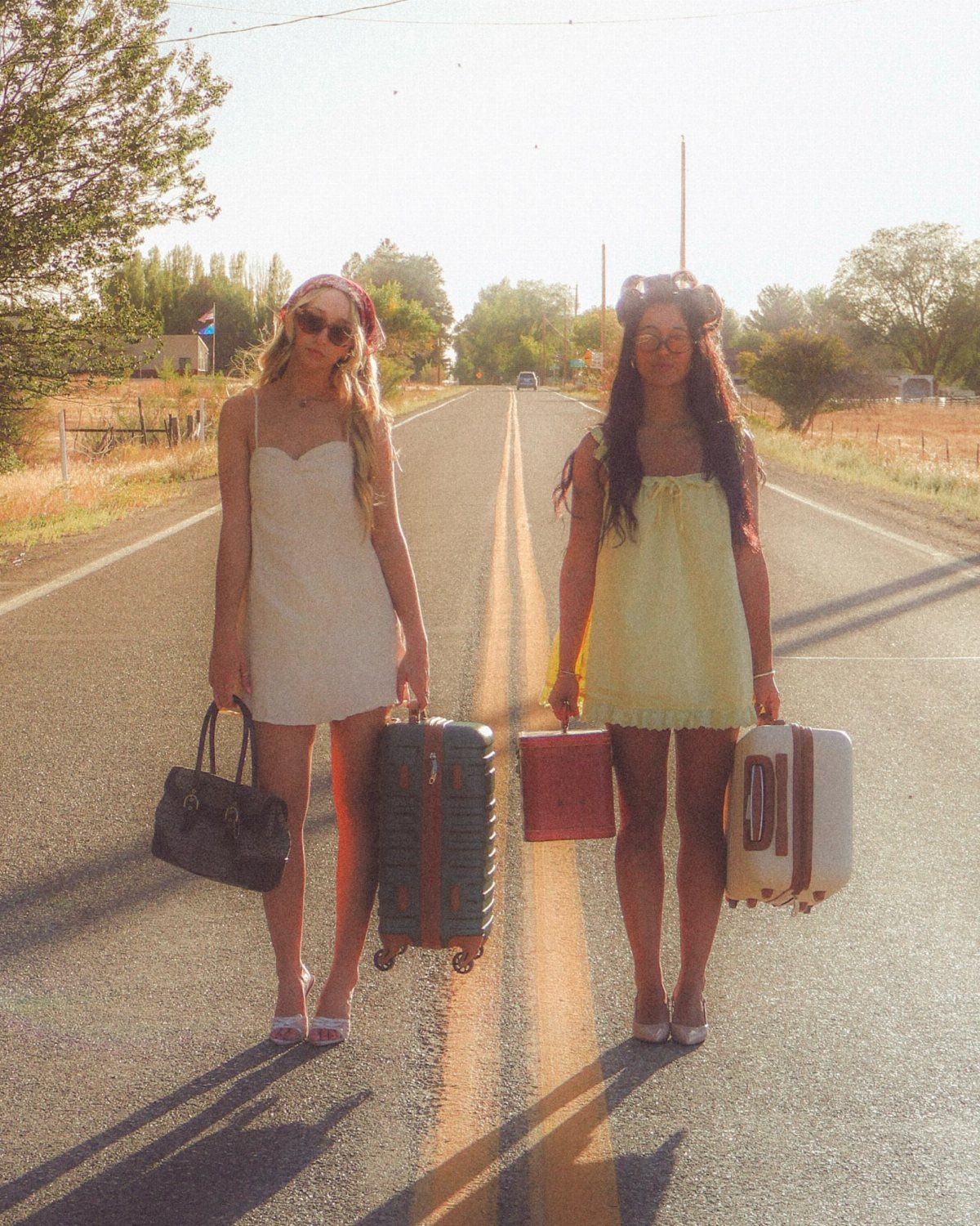 Two women in summer dresses standing on a road holding vintage suitcases.