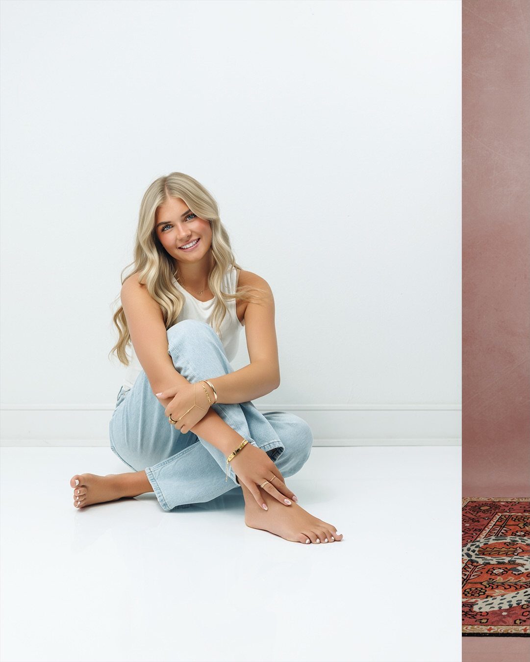 A smiling blonde woman sitting on a white floor wearing a white top and blue jeans.