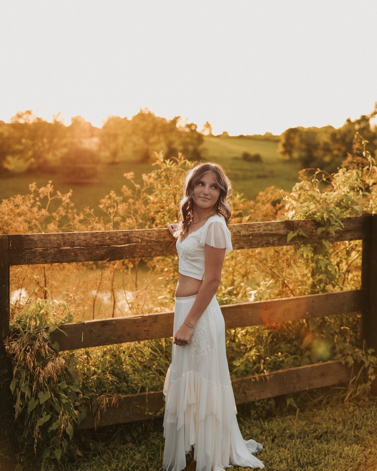 Young woman in a white lace crop top and long skirt standing by a wooden fence.
