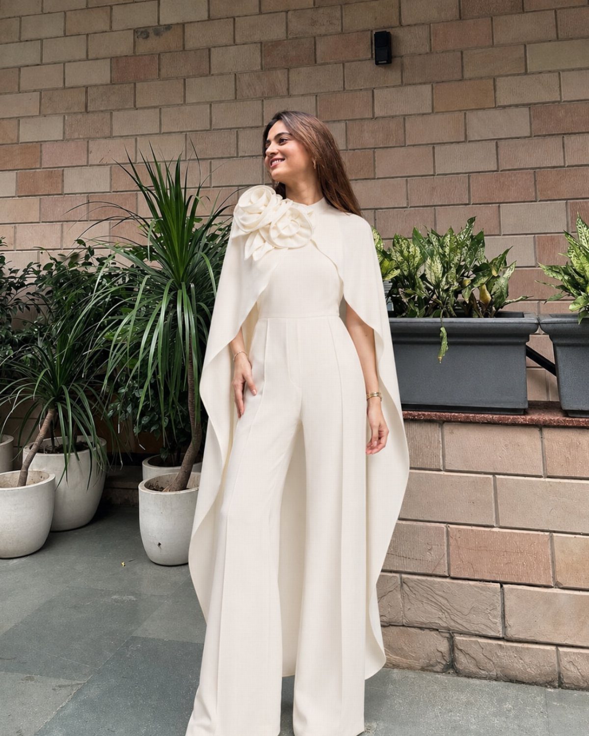 Woman smiling in a white wide-leg jumpsuit with cape sleeves and a floral shoulder detail.