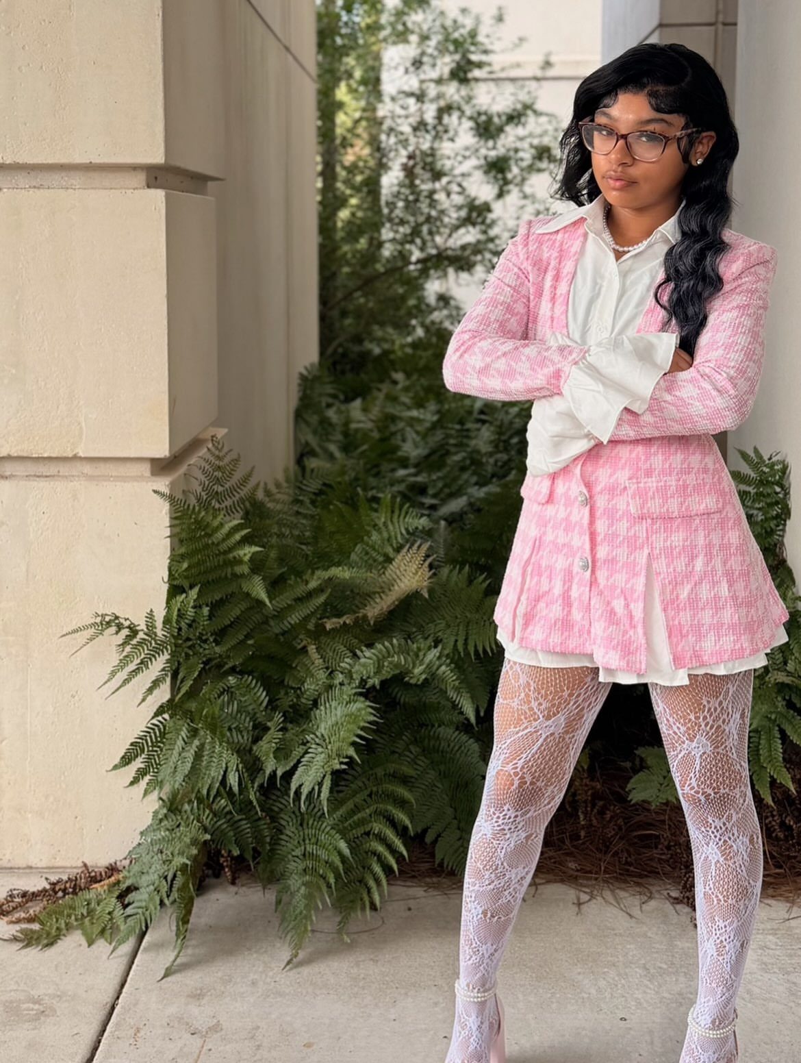A young woman wearing a pink patterned blazer dress and white patterned tights posing outdoors.