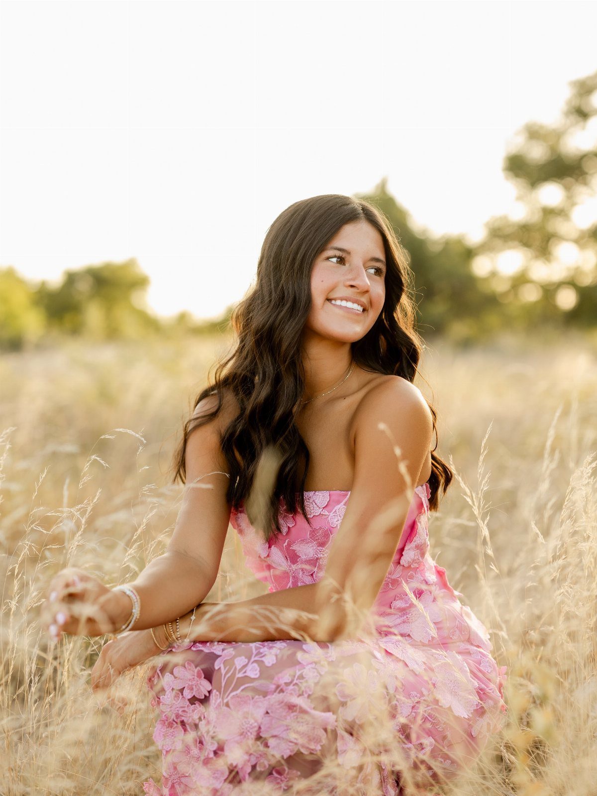 A smiling young woman sitting in a golden field wearing a pink lace dress.