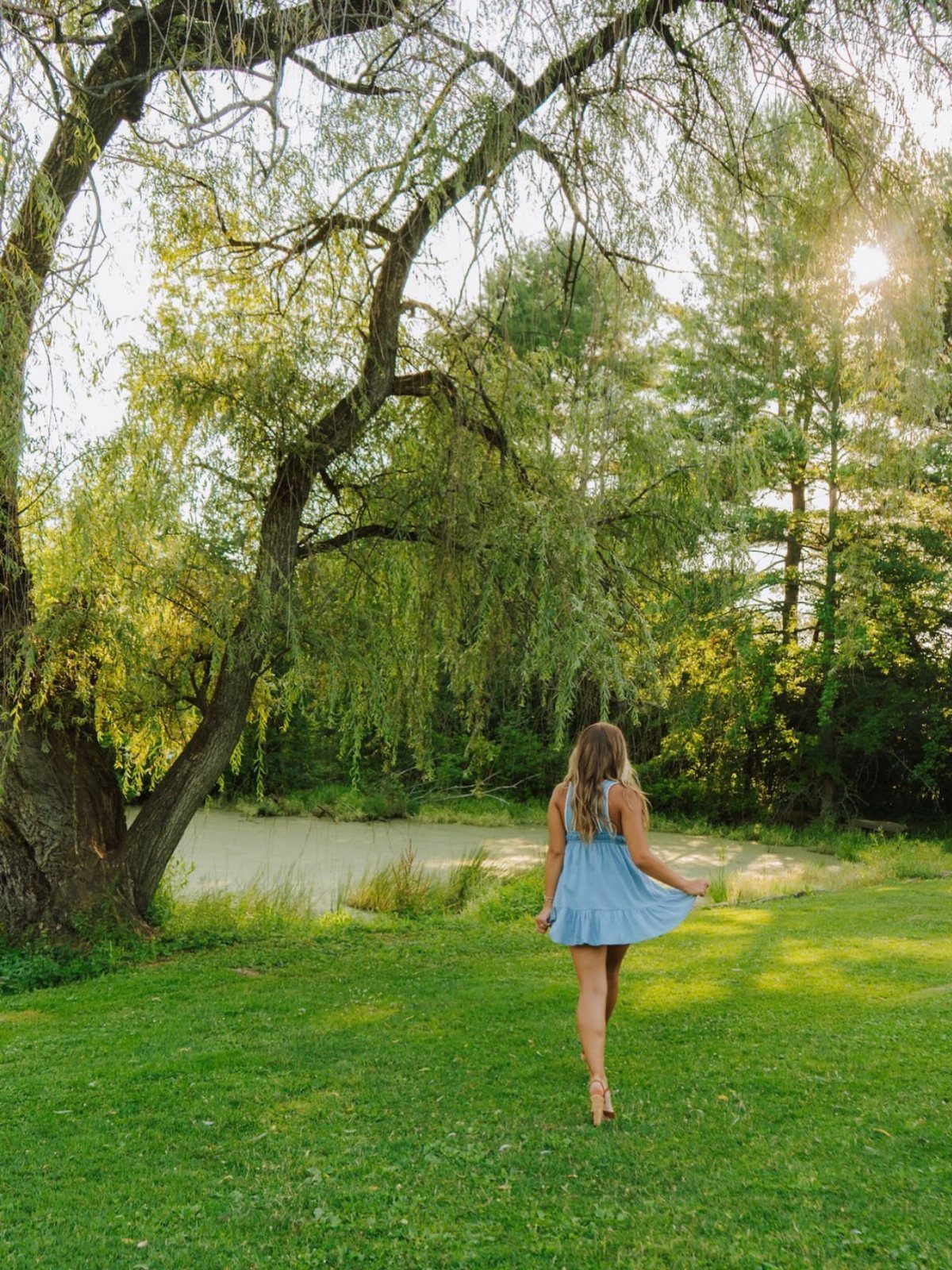 A young woman in a light blue dress walking through a sunny grassy field near trees.