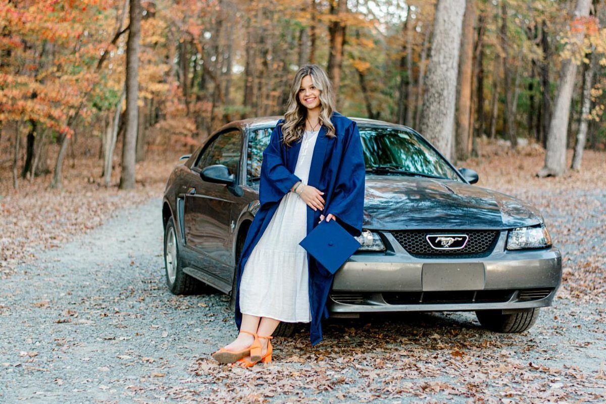 A smiling young woman in a white dress and blue graduation gown leaning against a car.