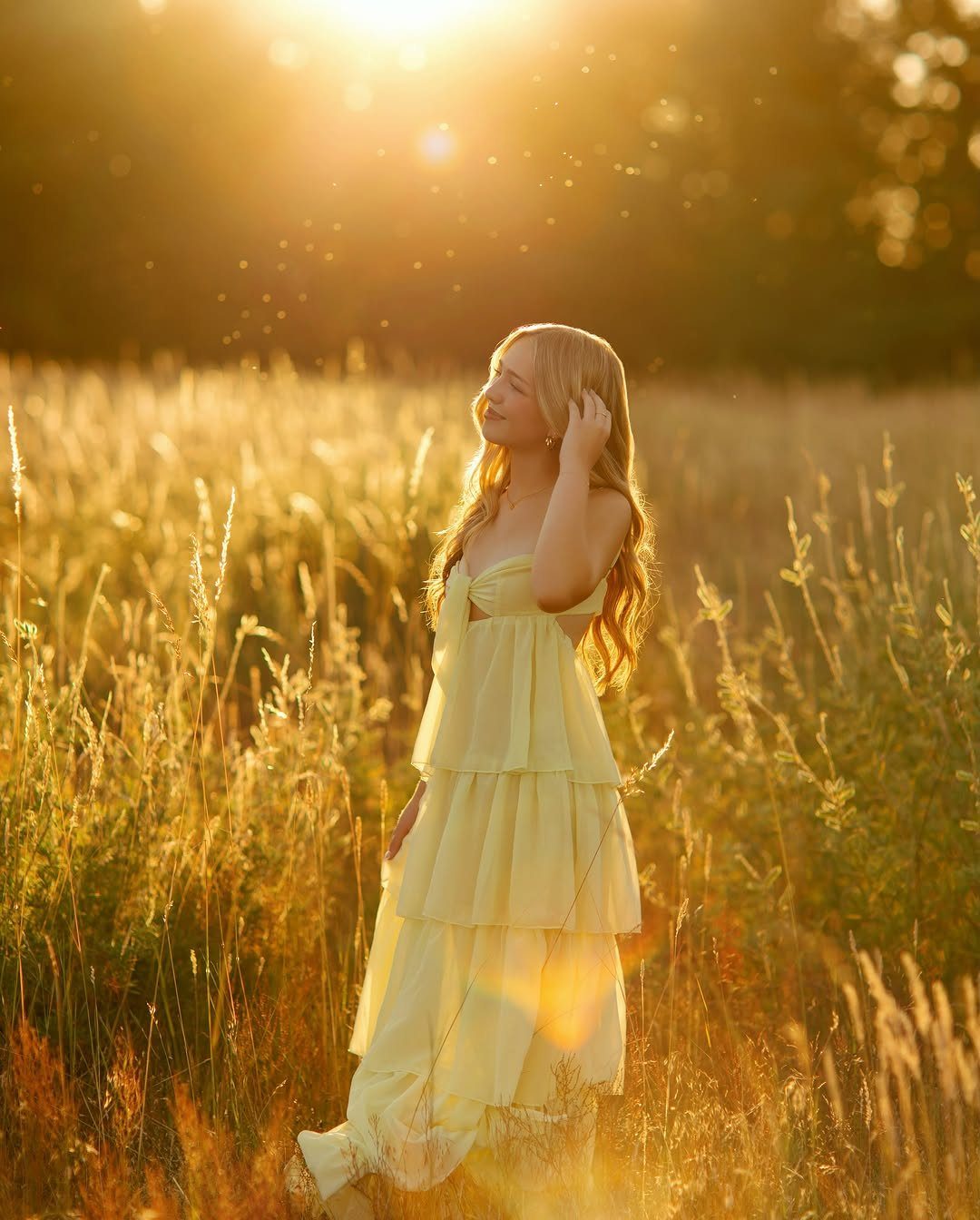 A young woman in a yellow tiered dress standing in a sunlit field of tall grass.