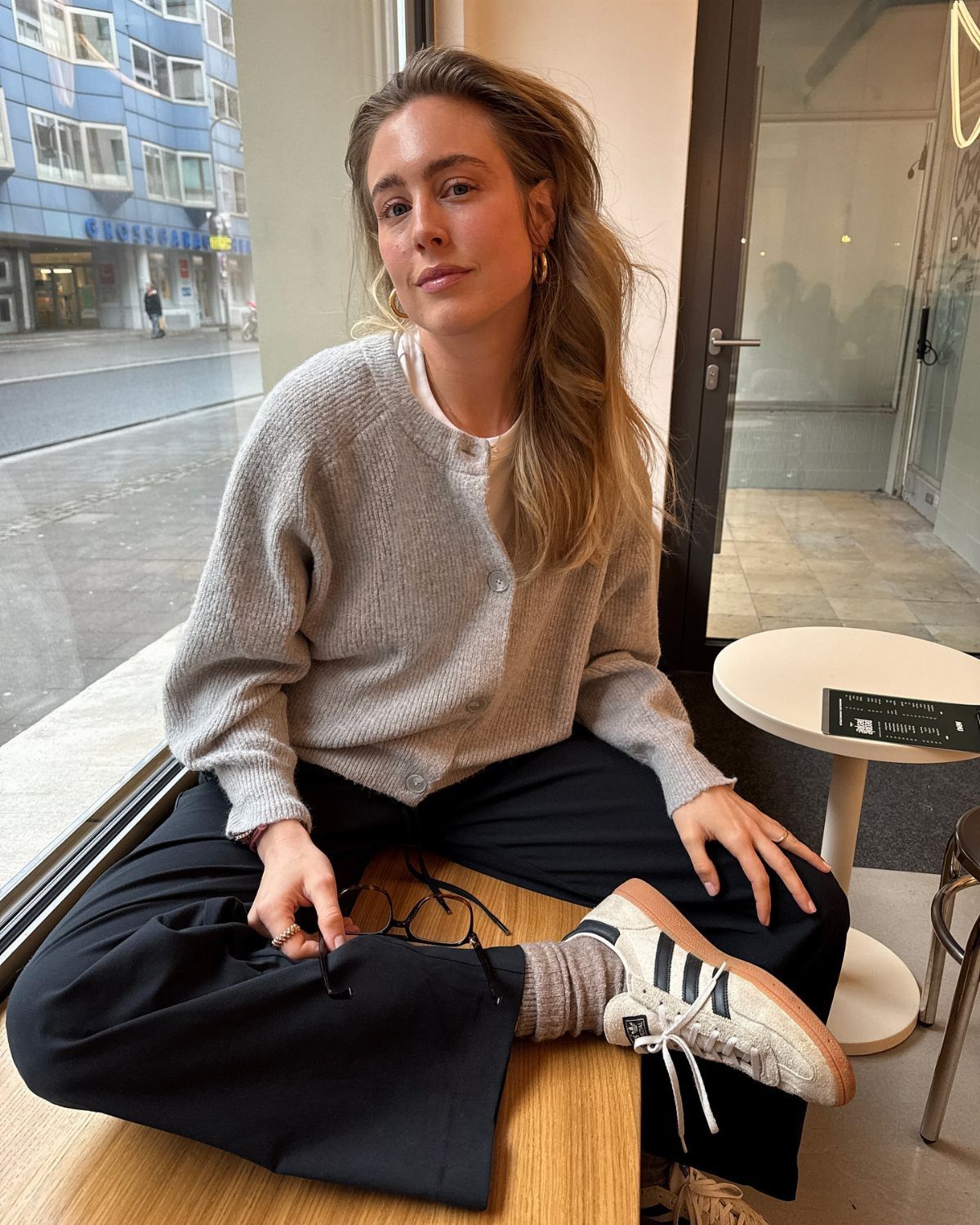 Woman wearing a grey knit cardigan, black trousers, and white sneakers sitting by a window.