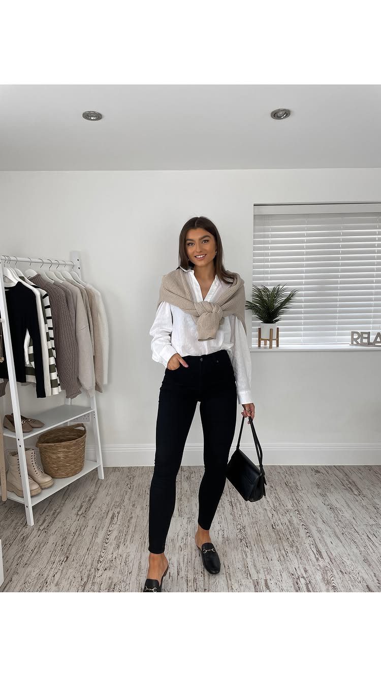 A woman posing in a white shirt, black jeans, and loafers with a sweater over her shoulders.
