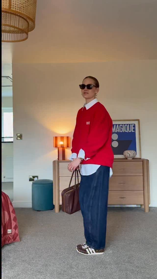 Woman wearing a red sweater layered over a white collared shirt with dark blue trousers.