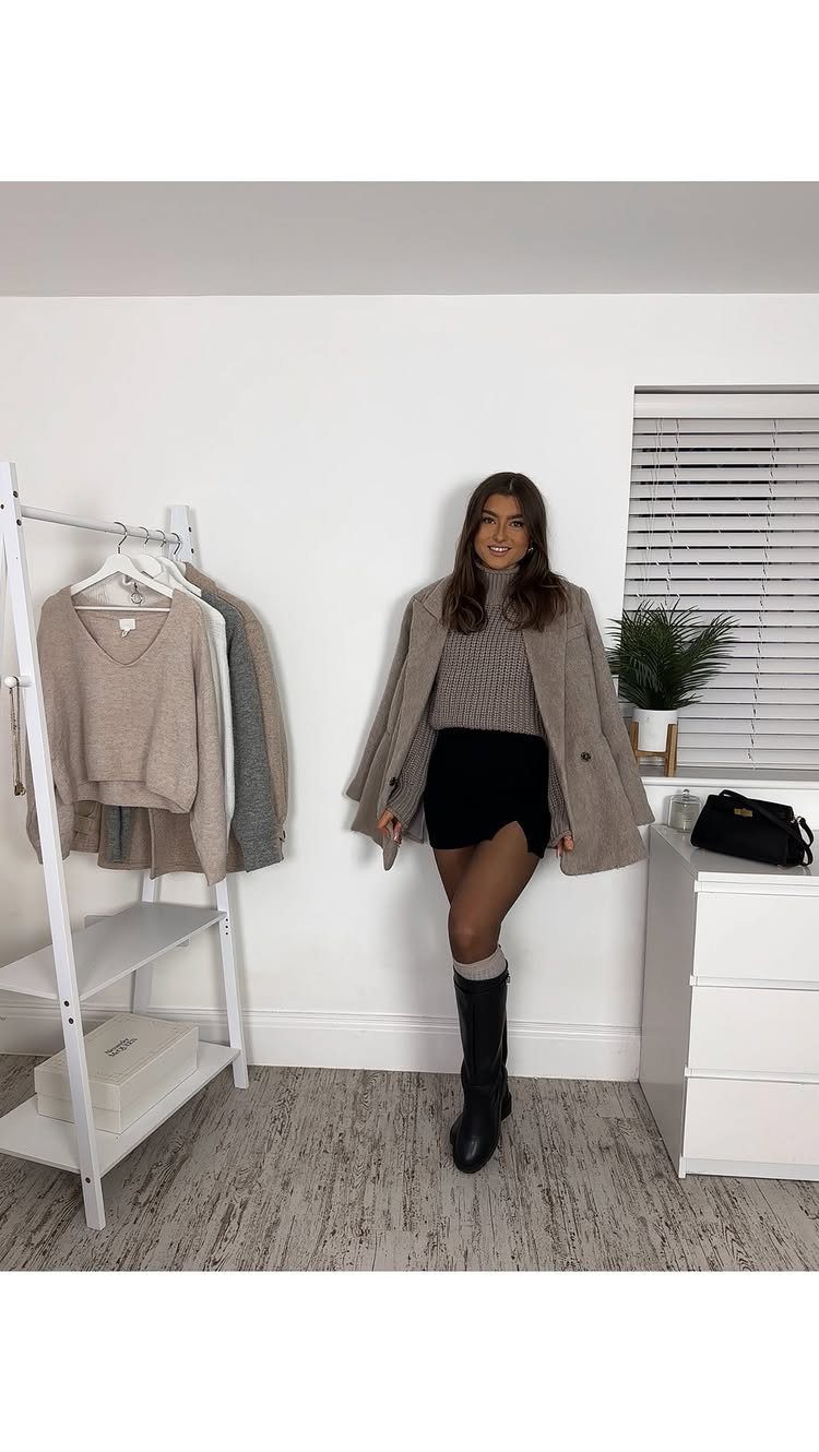 A woman wearing a beige sweater, black skirt, long coat, and black boots standing in a room.
