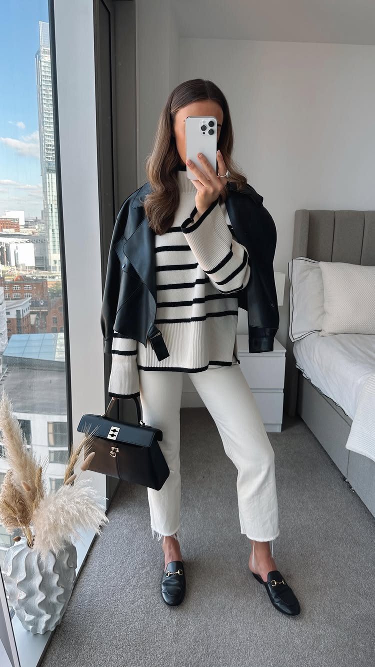 Woman in striped sweater, white trousers, and black leather jacket posing for a mirror selfie.